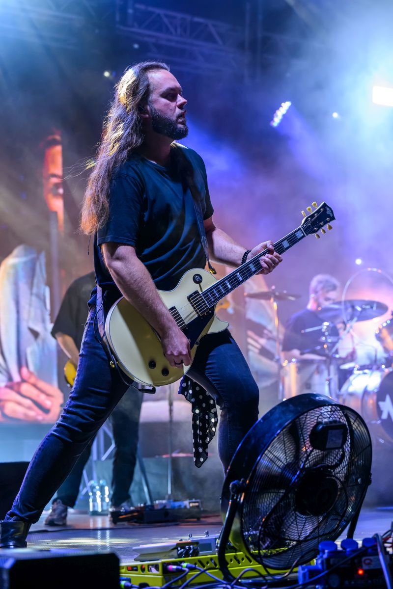 Mężczyzna z długimi włosami gra na kremowej gitarze na scenie. Tło rozświetlone efektami świetlnymi w odcieniach niebieskiego i fioletu. Przed nim znajduje się wentylator, a w tle widać perkusistę.