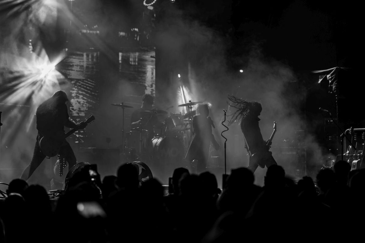 Na czarno-białym zdjęciu widzimy zespół rockowy na scenie w otoczeniu dymu. Muzycy z gitarami i perkusją, intensywne światła w tle. Publiczność z przodu w cieniu.