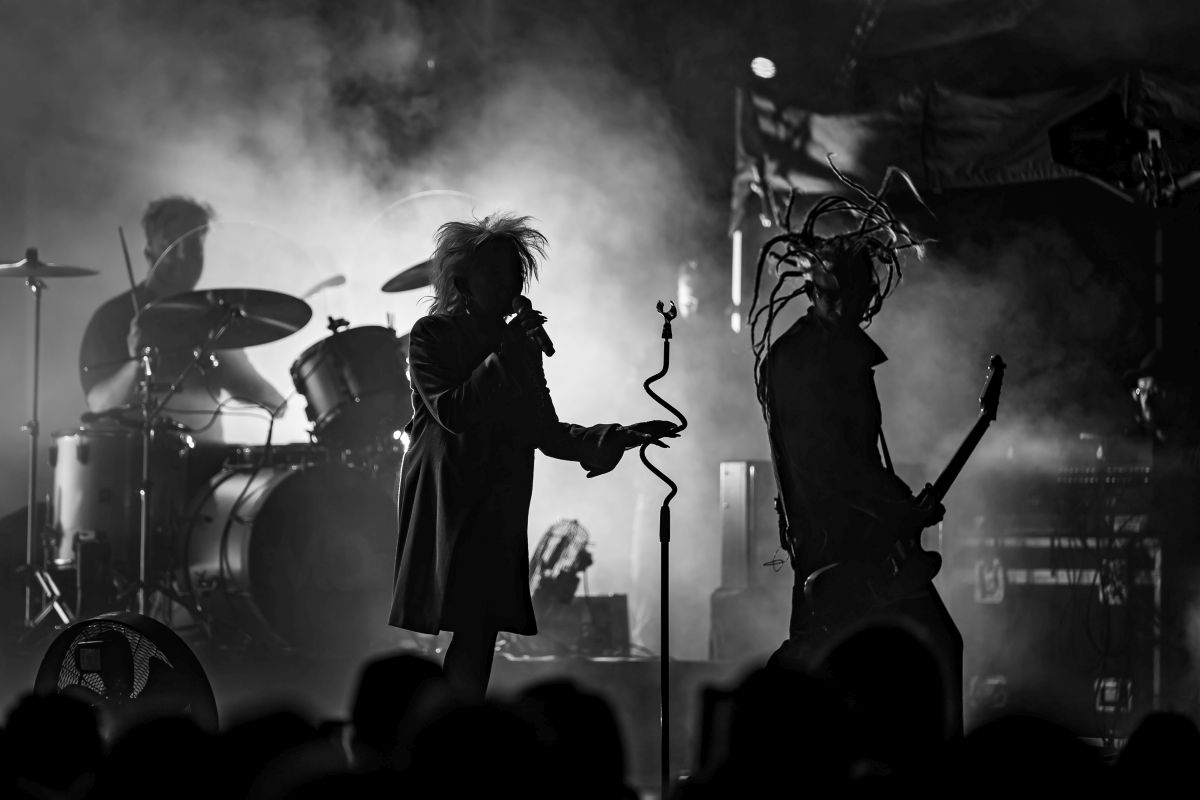 Na czarno-białym zdjęciu zespół rockowy na scenie. Wokalista z mikrofonem i basista z gitarą w dymie. Tło jest ciemne i rozmyte.