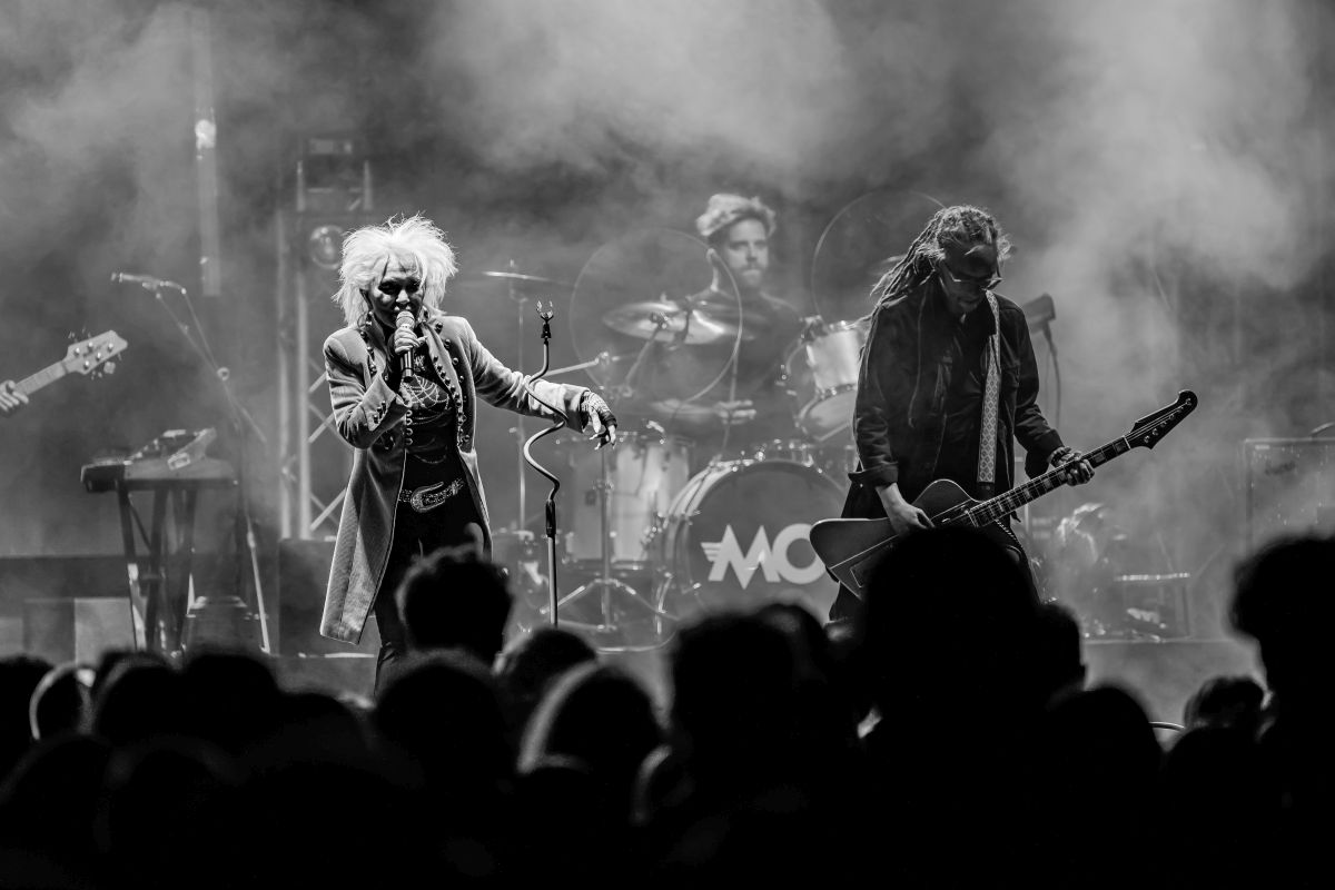Na czarno-białym zdjęciu grupa rockowa występuje na scenie. Wokalista trzyma mikrofon, gitarzysta z gitarą. W tle perkusista i publiczność.