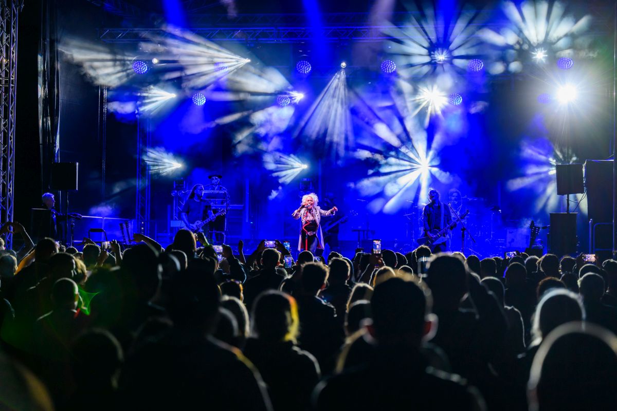 Koncert na żywo na scenie z niebieskimi światłami i dymem. Artysta w centrum, widownia z telefonami w górze. Tło w dynamice świateł.