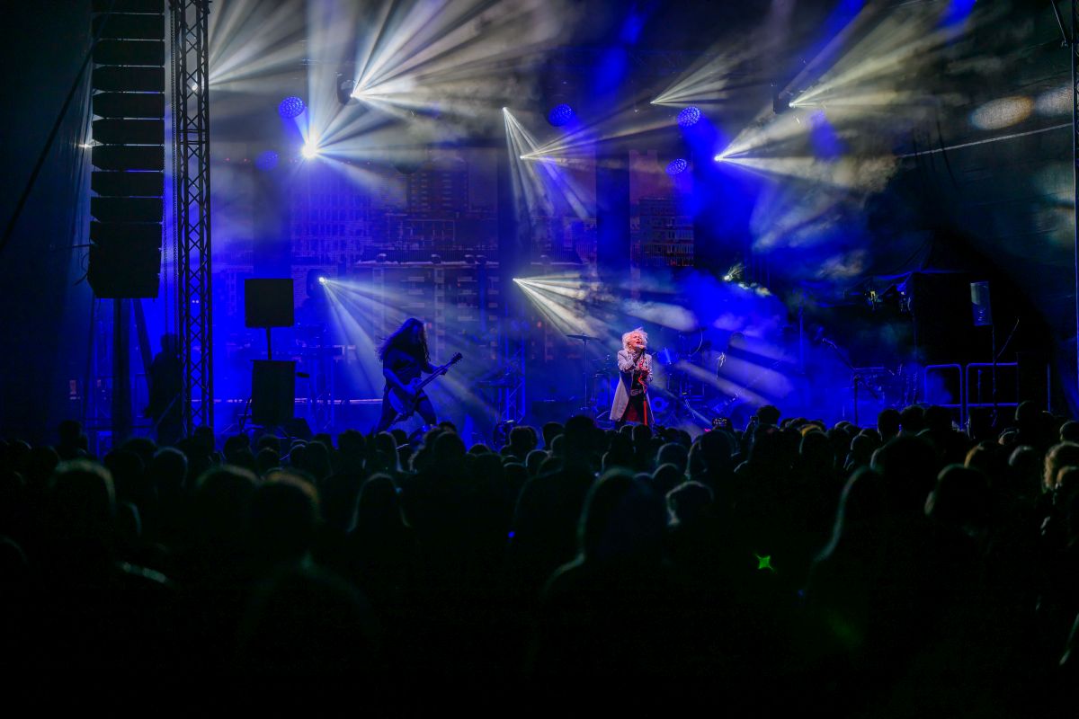 Koncert na scenie, oświetlonej niebieskimi i białymi światłami, z tłumem ludzi w ciemności. Gitarzysta po lewej, wokalista pośrodku. Dym tworzy dynamiczną atmosferę.