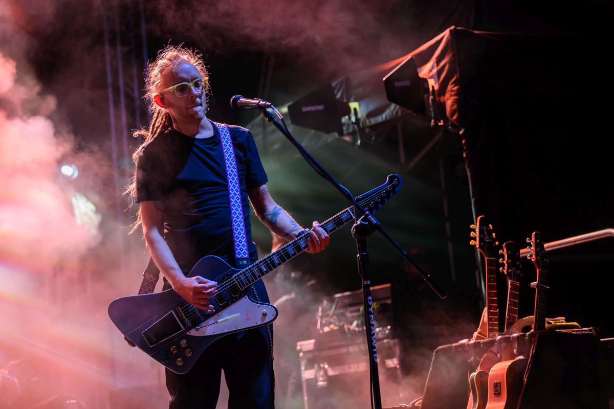 Muzyk gra na gitarze elektrycznej na scenie, w tle kolorowe światła i dym. Obok stoi kilka gitar. Dominuje ciemne, koncertowe oświetlenie.
