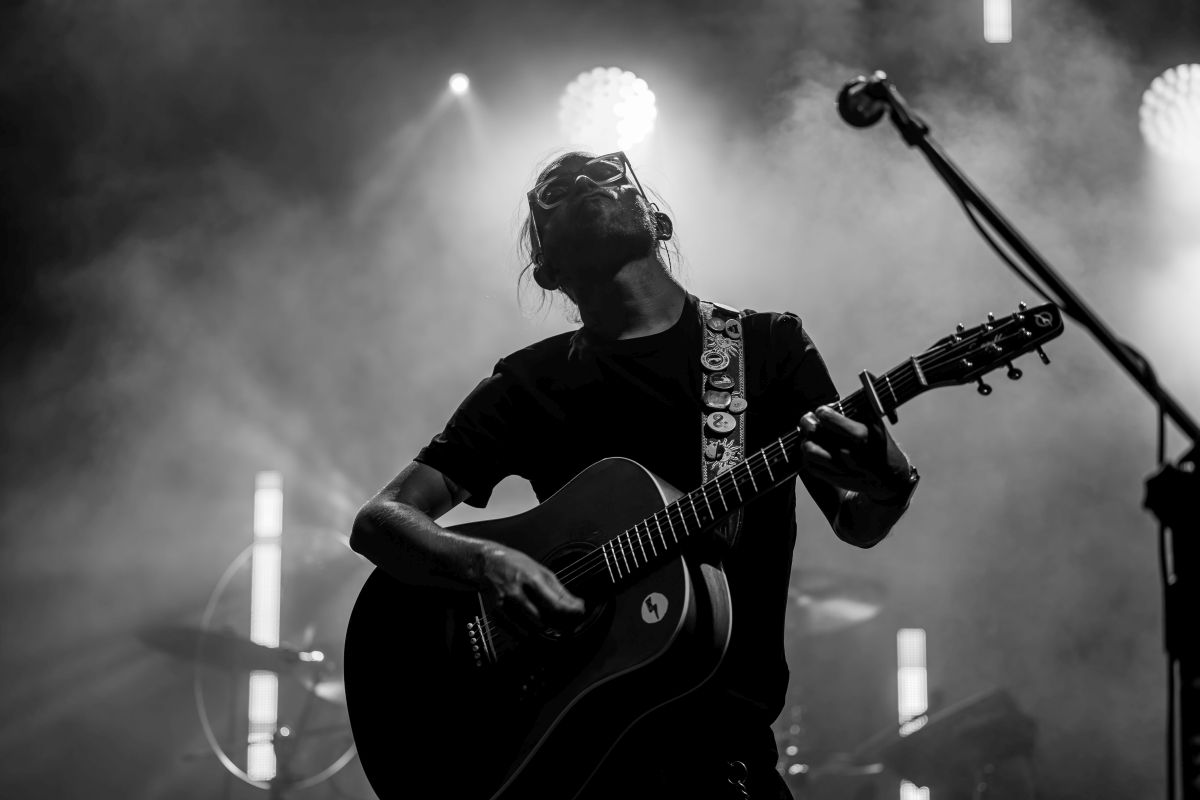 Muzyk gra na gitarze na scenie. Czarnobiałe zdjęcie pokazuje dym i światła w tle. Mikrofon widoczny z boku.