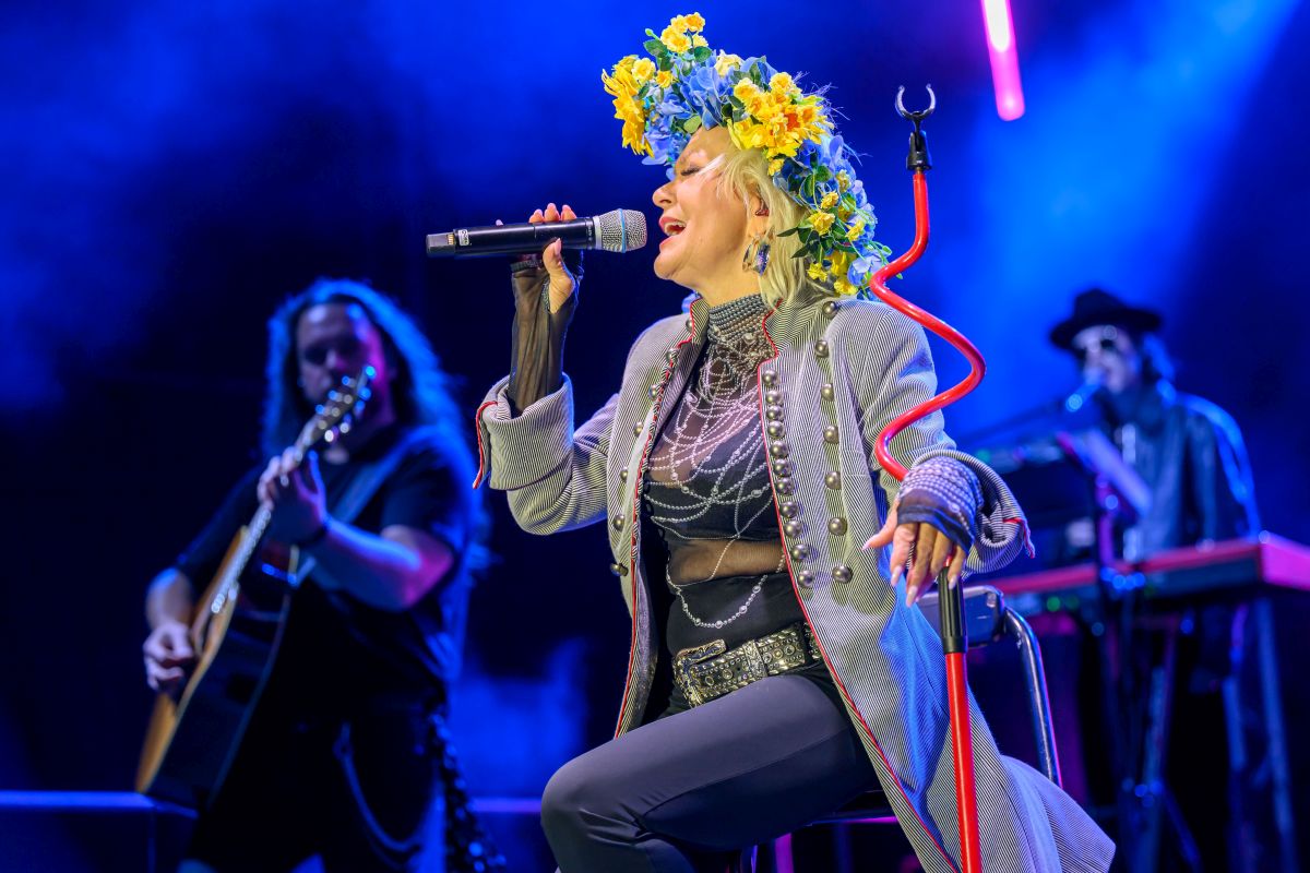 Wokalistka w wieńcu z kwiatów śpiewa na scenie. Za nią gitarzysta i klawiszowiec, scena oświetlona na niebiesko.