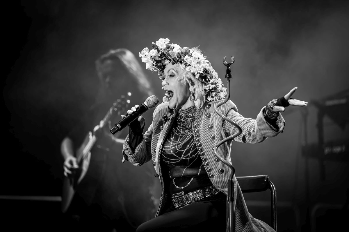 Na czarno-białym zdjęciu kobieta w kwiecistym wianku śpiewa na scenie. Nosi ozdobne ubranie z guzikami i koralikami. W tle widoczny muzyk z gitarą.