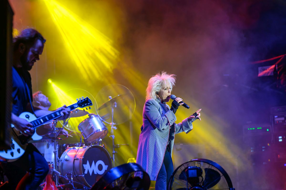 Na scenie zespół rockowy; piosenkarka w szarym płaszczu śpiewa, obok gitarzysta. Jasne, żółte światła oświetlają scenę. Tło w ciemnych barwach.