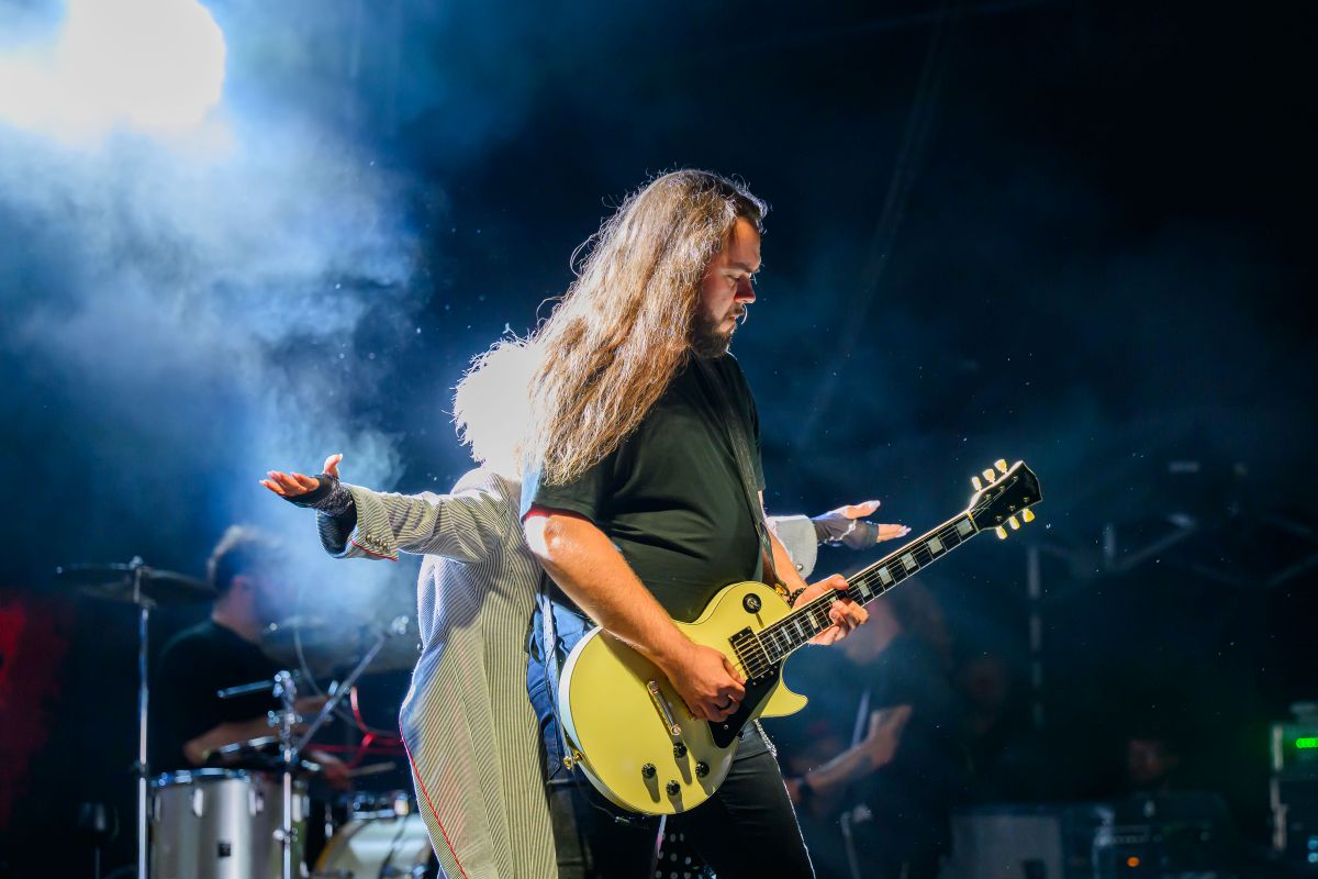 Na zdjęciu widać gitarzystę z długimi włosami na scenie, trzyma białą gitarę. Obok niego stoi osoba w jasnym stroju, z wyciągniętymi rękami. Tło jest ciemne, oświetlone niebieskim światłem i mgłą.