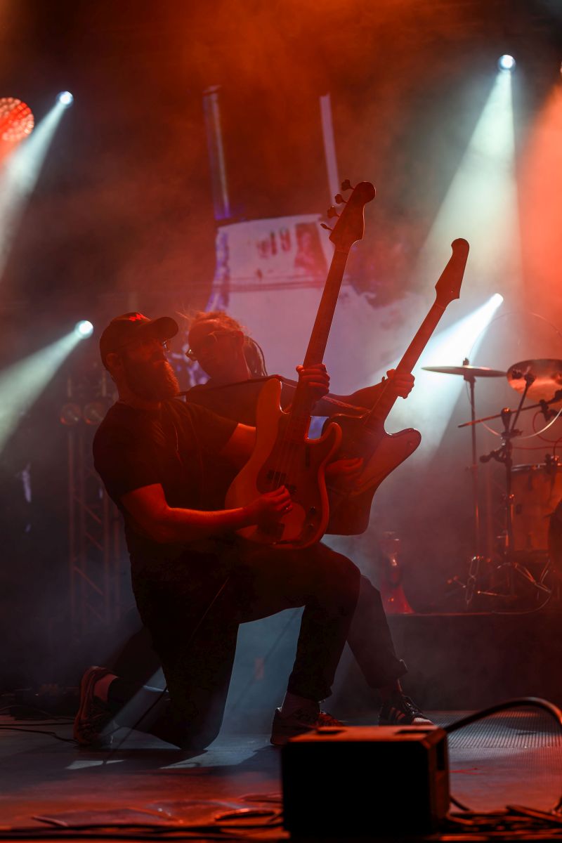 Na scenie dwóch muzyków gra na gitarach elektrycznych. Tło jest pełne intensywnego, kolorowego oświetlenia. Widać intensywnie czerwone i pomarańczowe reflektory oraz dym.