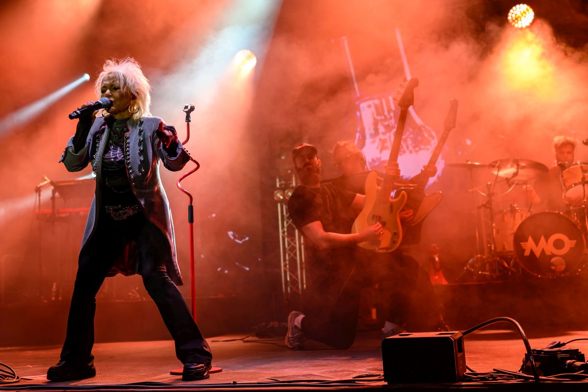 Na scenie kobieta śpiewa, trzymając mikrofon. Kolory są czerwono-pomarańczowe z efektami świetlnymi. W tle muzycy grają na gitarze i perkusji.