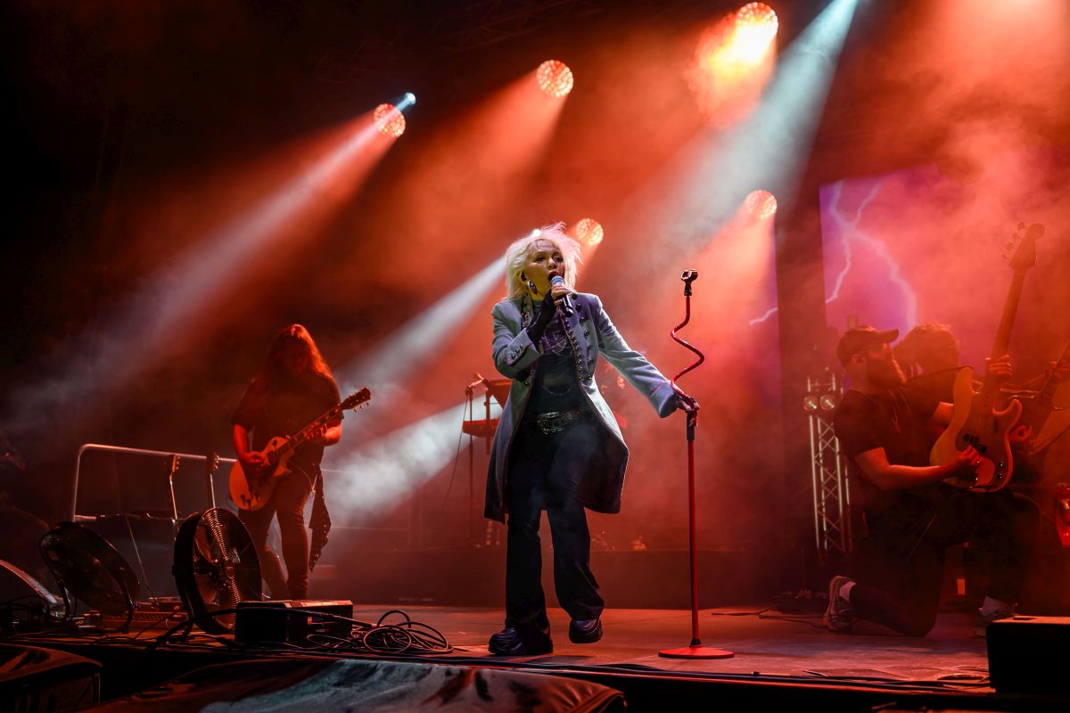 Na scenie muzyk w jasnym płaszczu śpiewa przy mikrofonie. Czerwone i białe światła oświetlają scenę. W tle dym i grający gitarzyści.