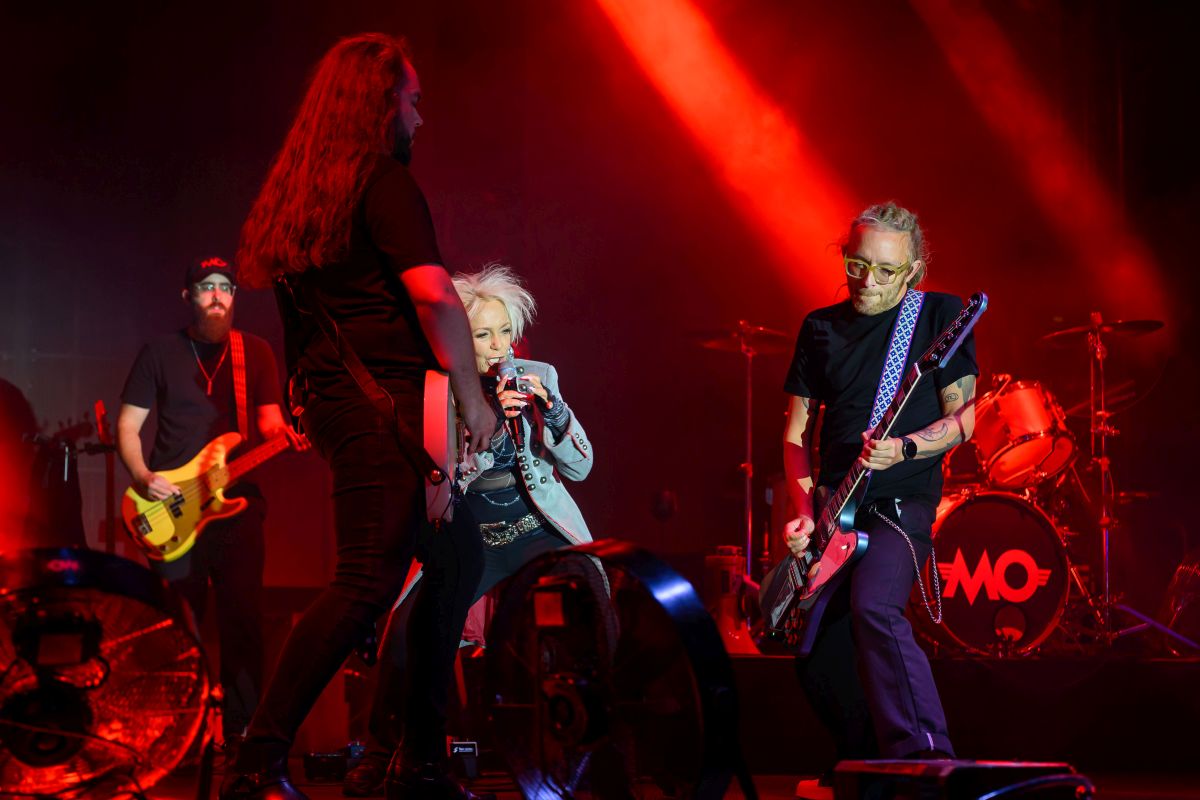 Na scenie czwórka muzyków grających rocka, w tle zestaw perkusyjny w czerwonych światłach. Gitarzysta z brodą i basista z długimi włosami. Wokalistka w szarej kurtce śpiewa do mikrofonu.