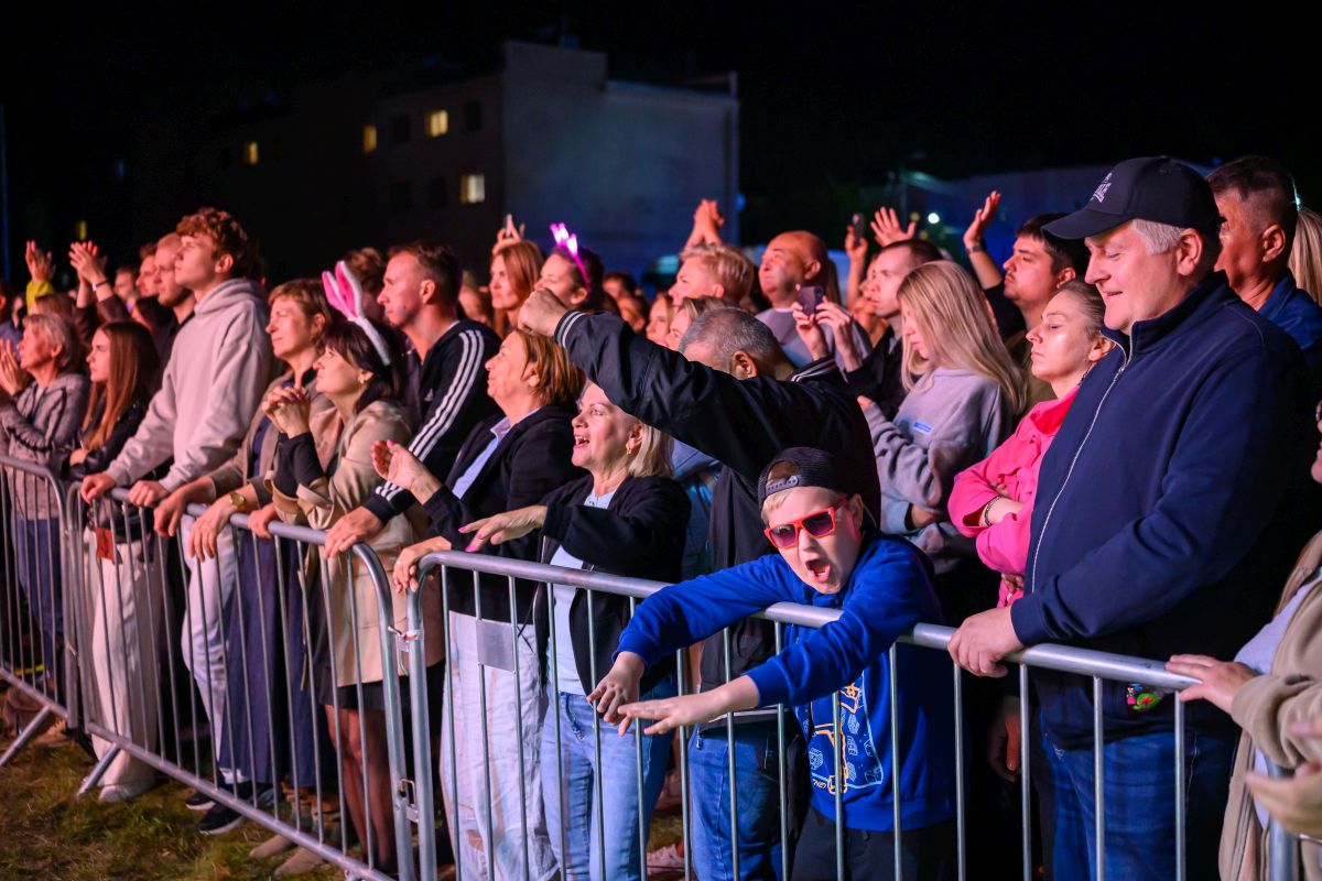 Grupa ludzi ogląda koncert za barierką nocą. Tłum jest różnorodny, widać jasno oświetlone twarze. Chłopiec w niebieskiej bluzie i czerwonych okularach wyróżnia się na pierwszym planie.