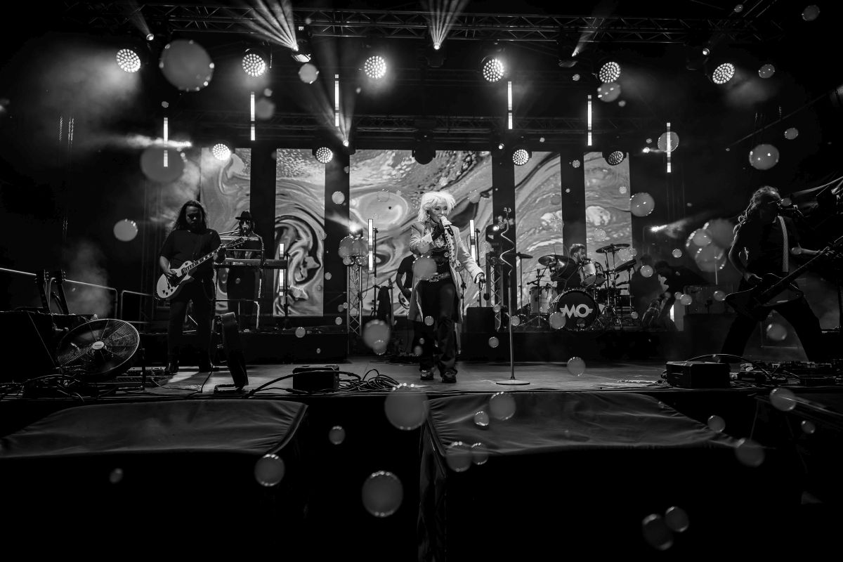 Czarno-białe zdjęcie zespołu na scenie z instrumentami, światła padają na muzyków. W tle abstrakcyjne wizualizacje. Unoszą się bańki.