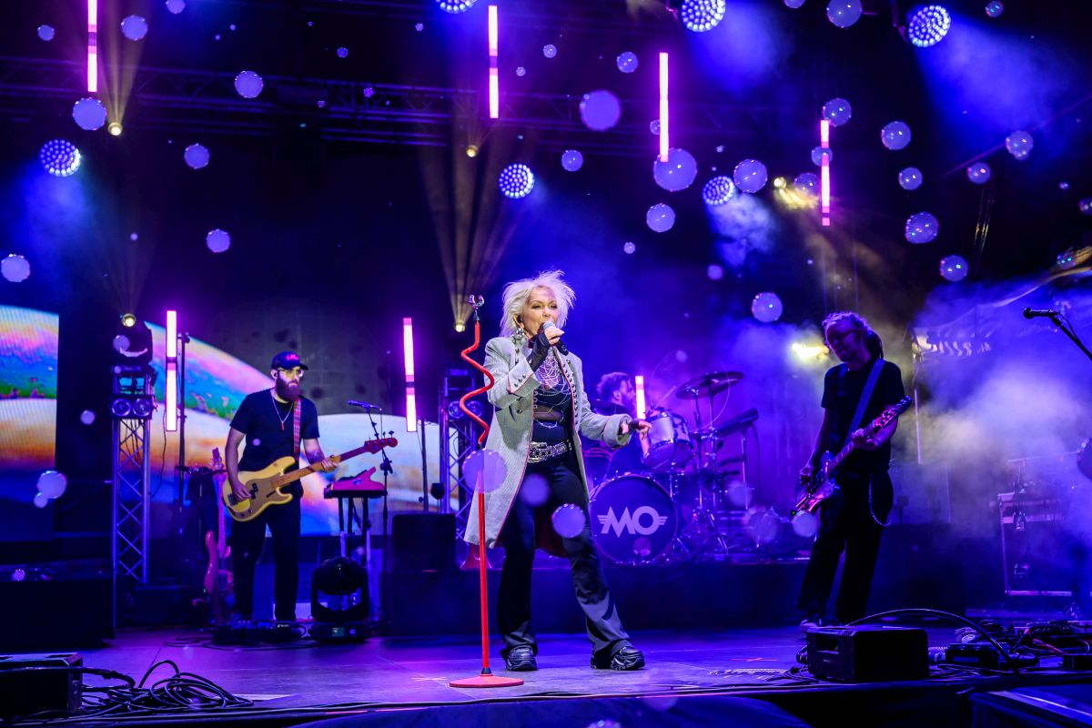 Na scenie zespół gra koncert, otoczony fioletowo-niebieskimi światłami. Wokalista śpiewa, trzymając mikrofon. Tło składa się z kolorowych świateł i efektów dymu.
