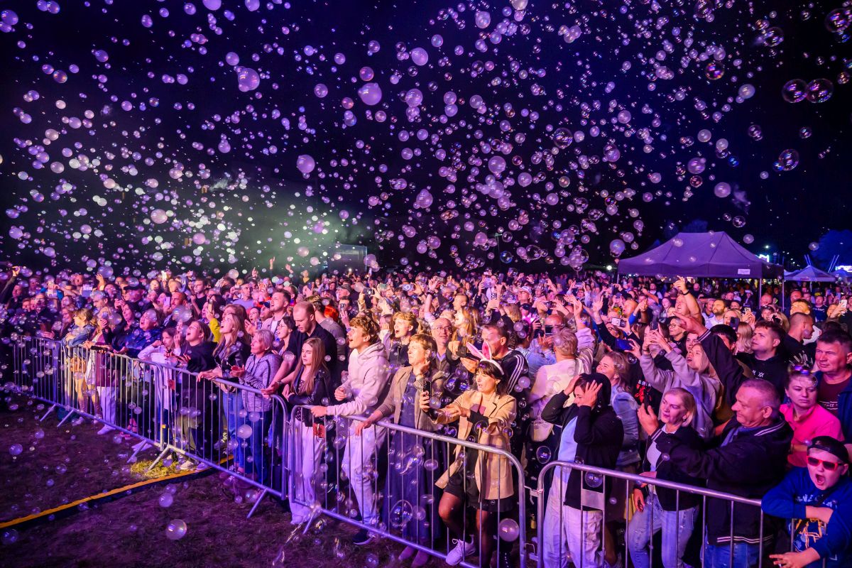 Tłum zgromadzony na koncercie w nocy z milionem mydlanych baniek. Ludzie za metalowym ogrodzeniem są podekscytowani. Światła sceniczne tworzą kolorowe iluminacje.