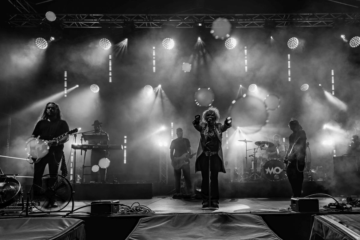 Czarno-białe zdjęcie przedstawia zespół muzyczny na scenie. W tle widać intensywne światła i dym. Muzycy grają na gitarach, perkusji i klawiszach.