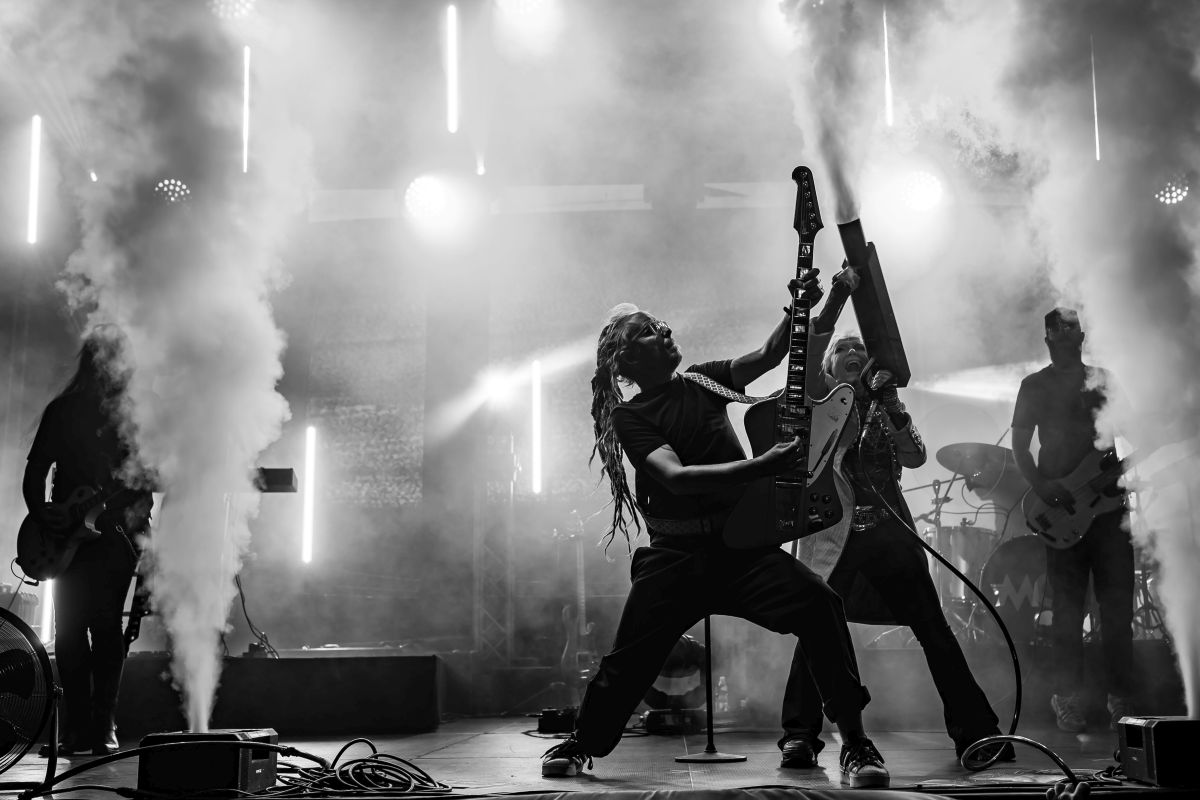 Zespół rockowy na scenie w dynamicznej pozie. Czarnobiałe zdjęcie z efektami dymu i jasnym oświetleniem w tle. Muzycy z gitarami, pełni energii.