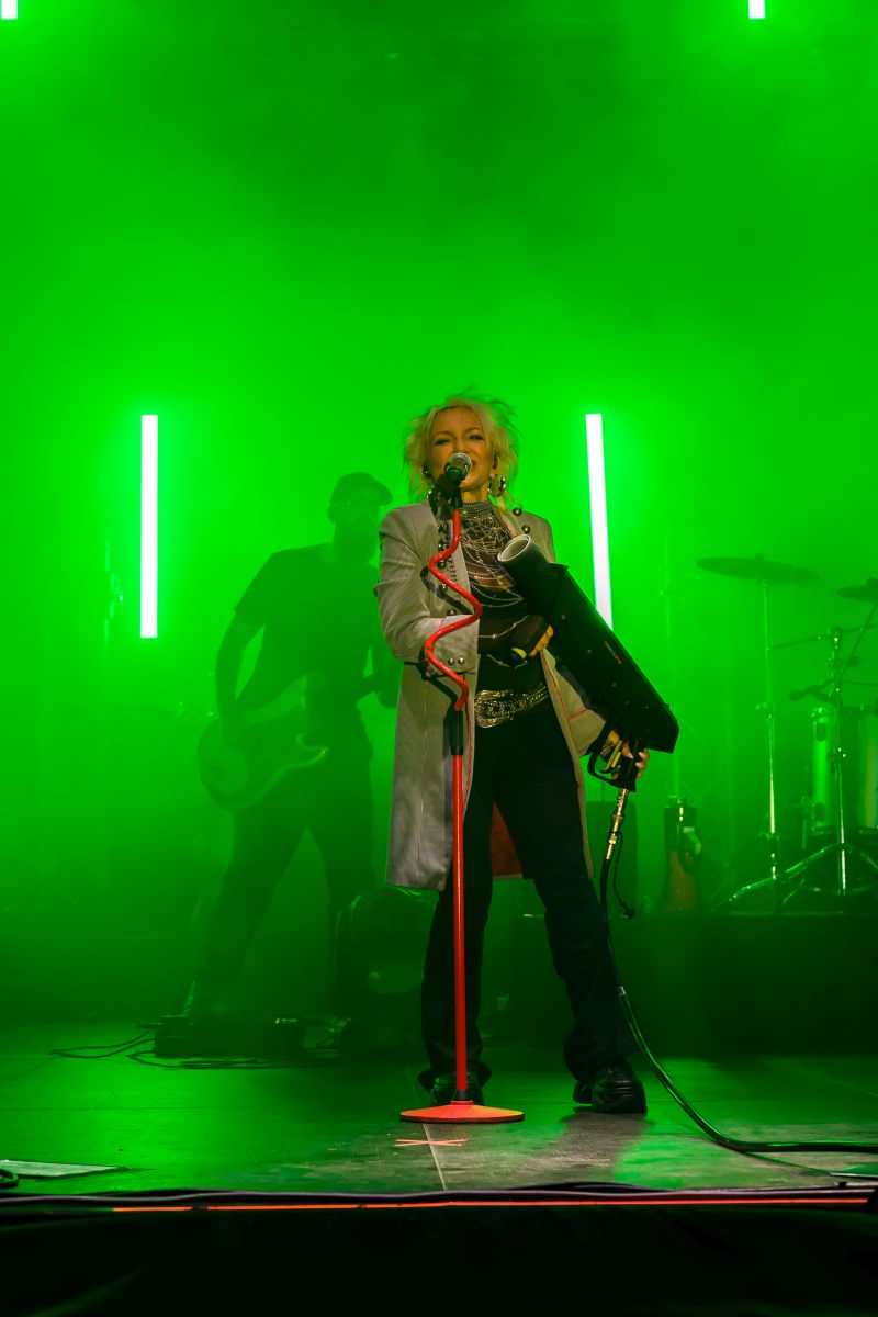 Osoba śpiewa na scenie z zielonym oświetleniem. Tło jest dymne, widoczny jest gitarzysta i perkusja. Scena ma intensywny, futurystyczny klimat.