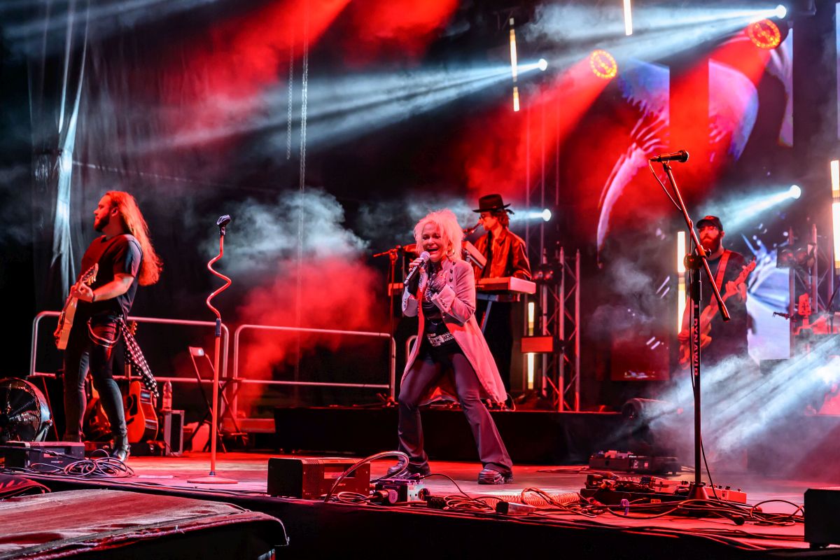 Koncert rockowy z zespołem na scenie, oświetlenie w czerwonych i niebieskich barwach. Wokalista w centrum z mikrofonem. W tle instrumenty i dym.
