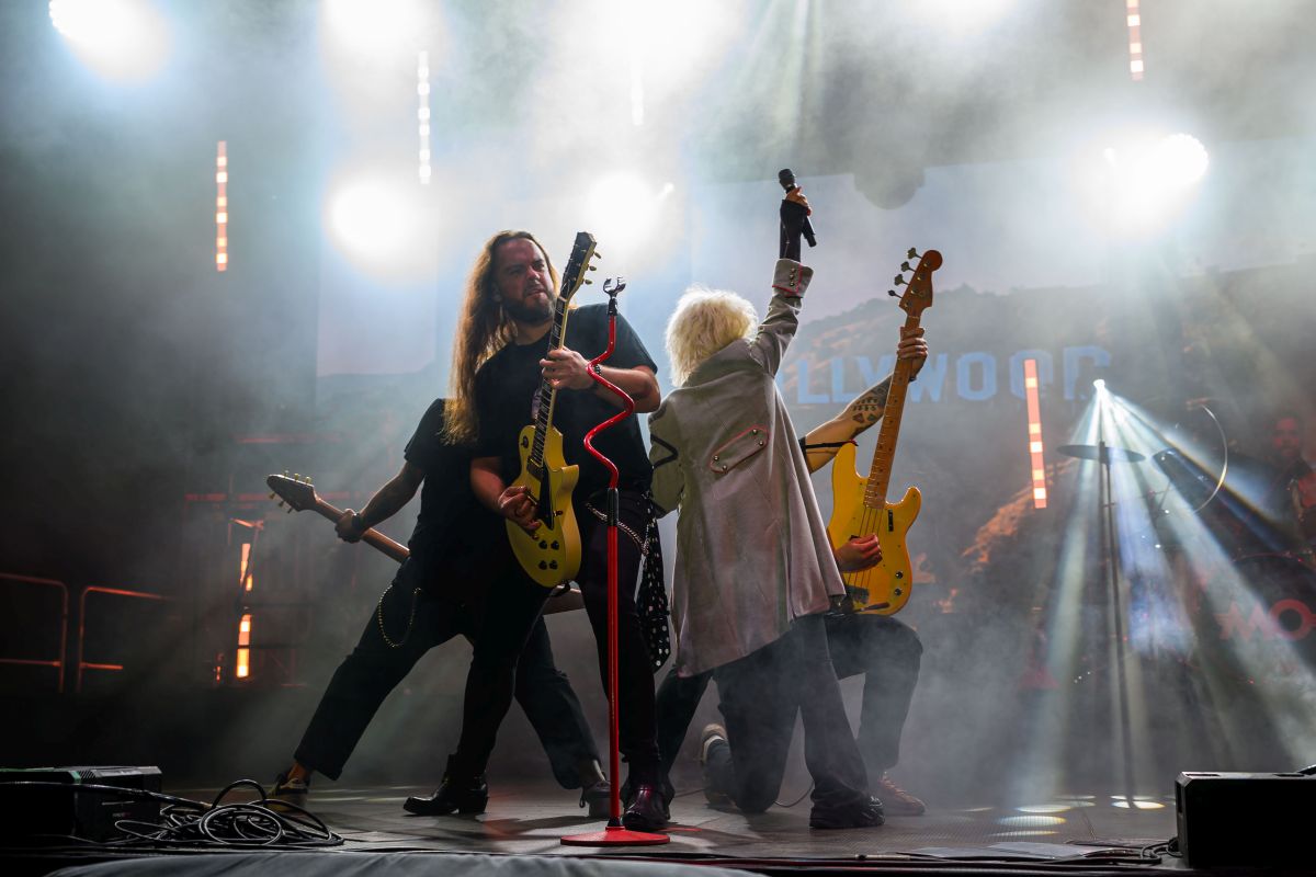 Zespół rockowy na scenie w jasnych światłach. Dwóch gitarzystów i wokalista z podniesionym mikrofonem na tle napisu Hollywood. Kolory dominujące to czerń i żółty.