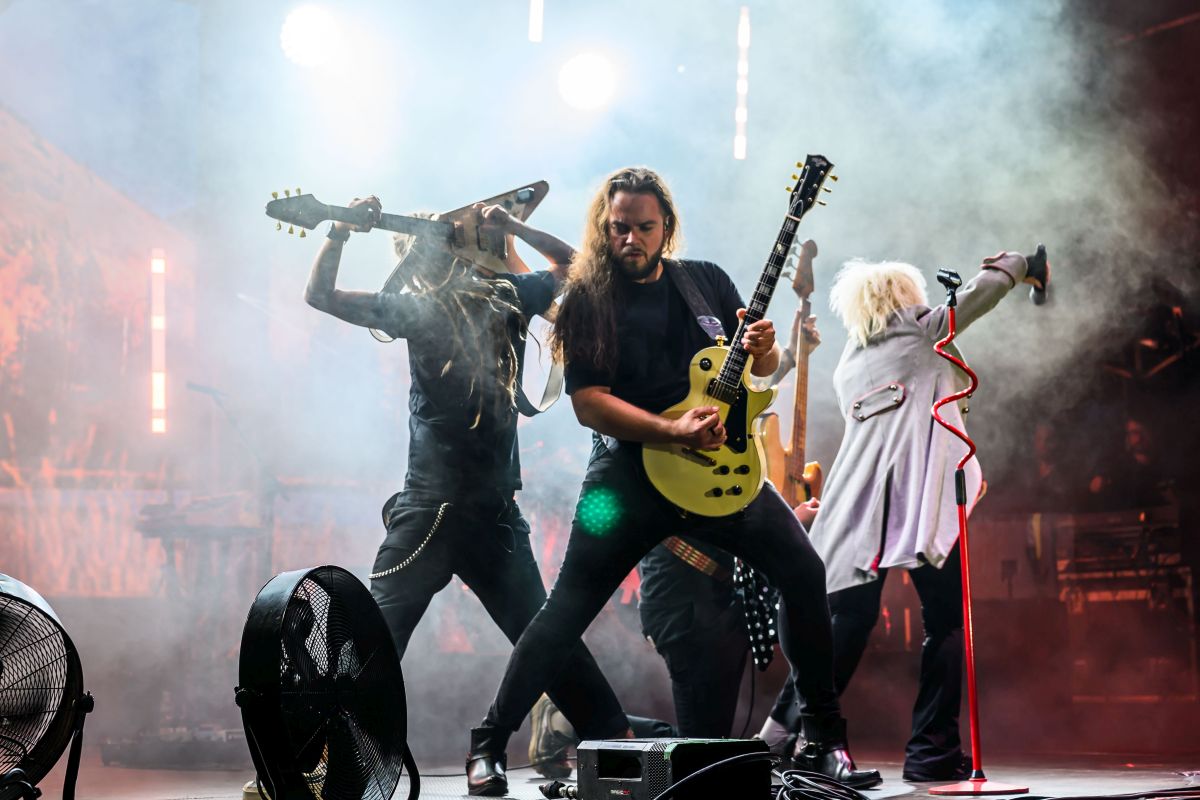 Trzech muzyków na scenie gra na gitarach. Scena oświetlona jest intensywnymi reflektorami, w tle unoszą się dymy. Centralnie mężczyzna z żółtą gitarą.