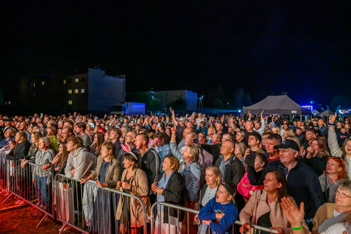 Tłum ludzi obserwuje koncert na świeżym powietrzu nocą. W tle ciemne niebo i namioty. Publiczność przy barierkach pełna emocji.