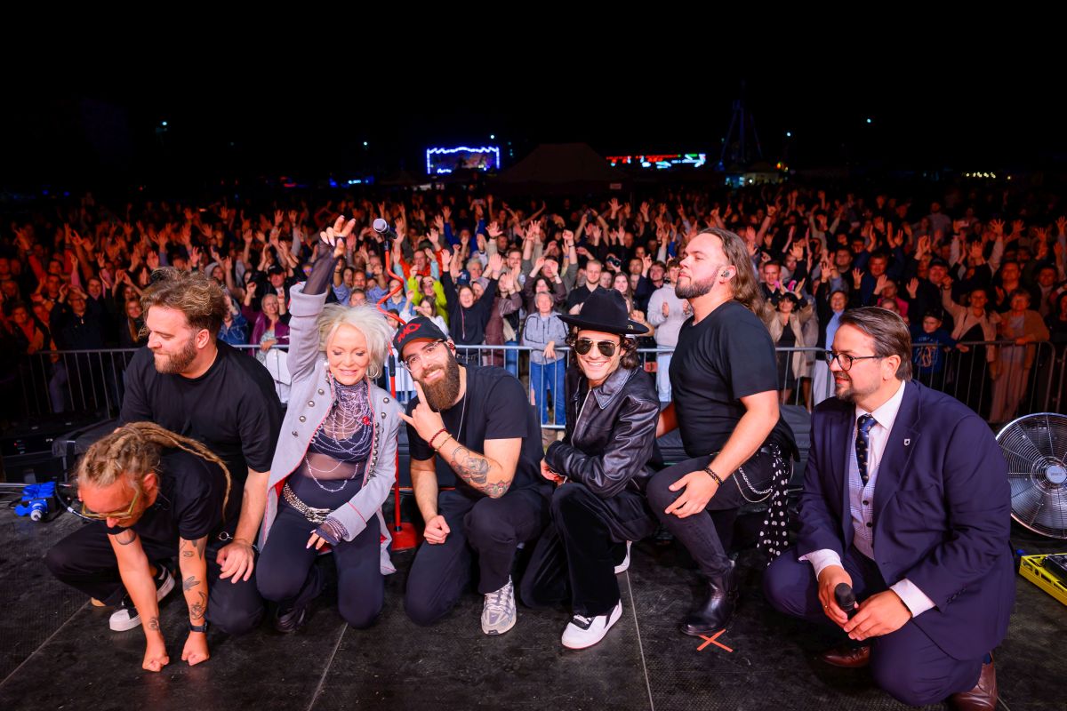 Grupa osób na scenie pozuje do zdjęcia z tłumem ludzi w tle. Jasne światła oświetlają scenę, tworząc kontrast z ciemnym niebem. Wydarzenie ma charakter koncertu lub spotkania publicznego.