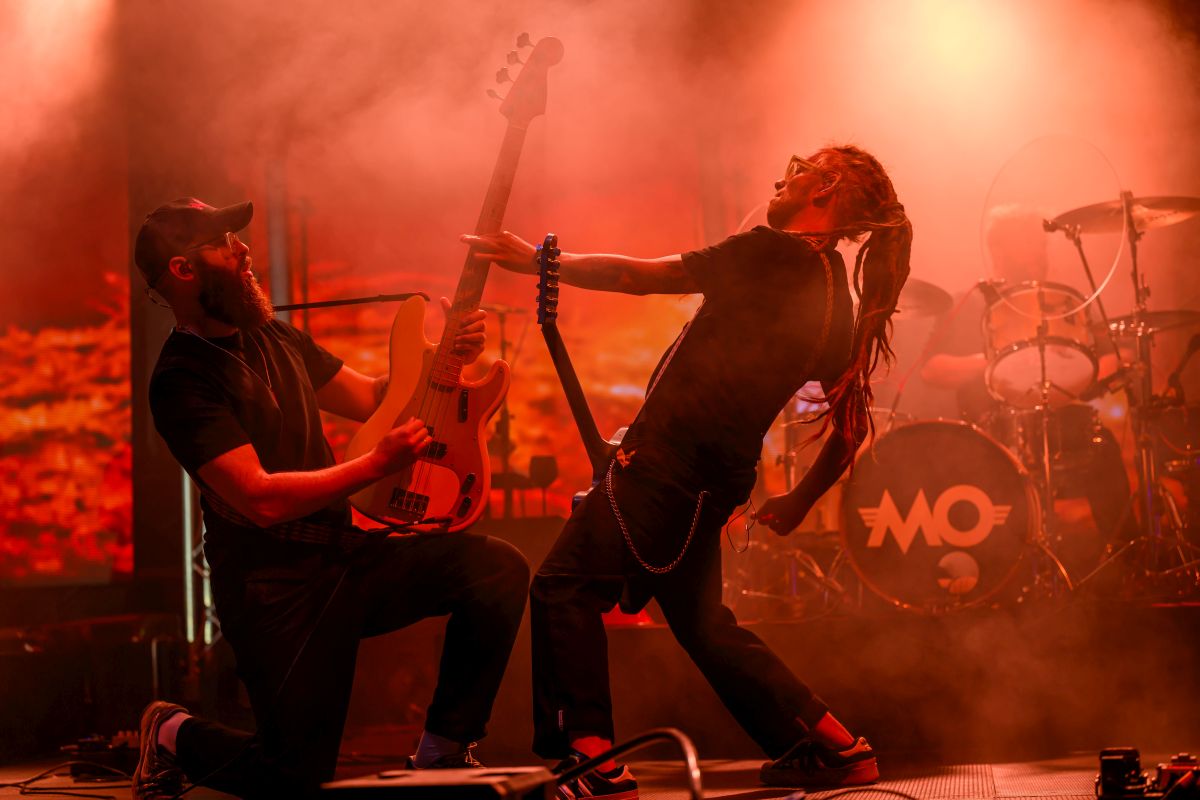 Dwóch muzyków na scenie z gitarą i perkusją, oświetlonych czerwonym światłem. W tle mocny dym. Napięta, energetyczna atmosfera.