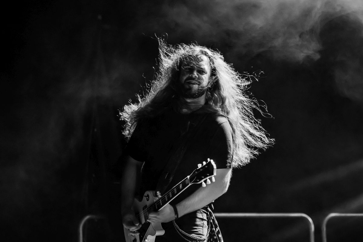 Muzyk z długimi włosami gra na gitarze elektrycznej na scenie. Czarno-białe zdjęcie z dynamicznym oświetleniem. Tło jest ciemne i zamglone.