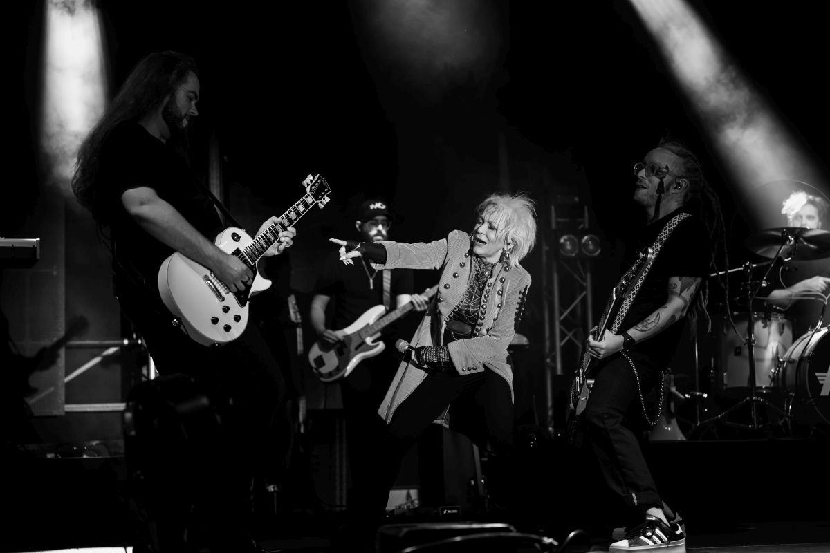 Na scenie zespół rockowy w czarno-białych barwach; gitarzysta, wokalistka i perkusista oświetleni reflektorami. Wokalistka dynamicznie gestykuluje w stronę gitarzysty. Tło zaciemnione, z widocznymi instrumentami i sprzętem koncertowym.
