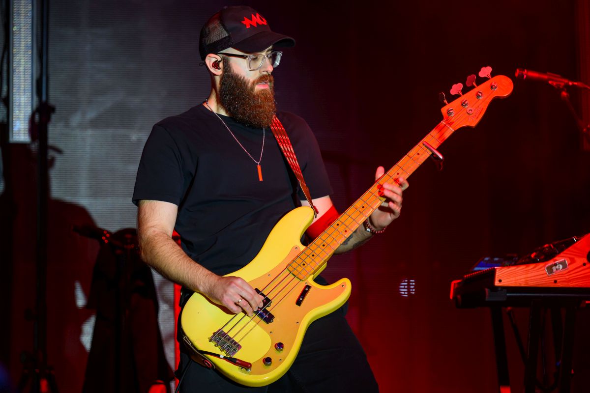 Muzyk z brodą gra na żółtej gitarze basowej na scenie. Nosi czarną czapkę i okulary. Tło jest ciemne z czerwonym oświetleniem.