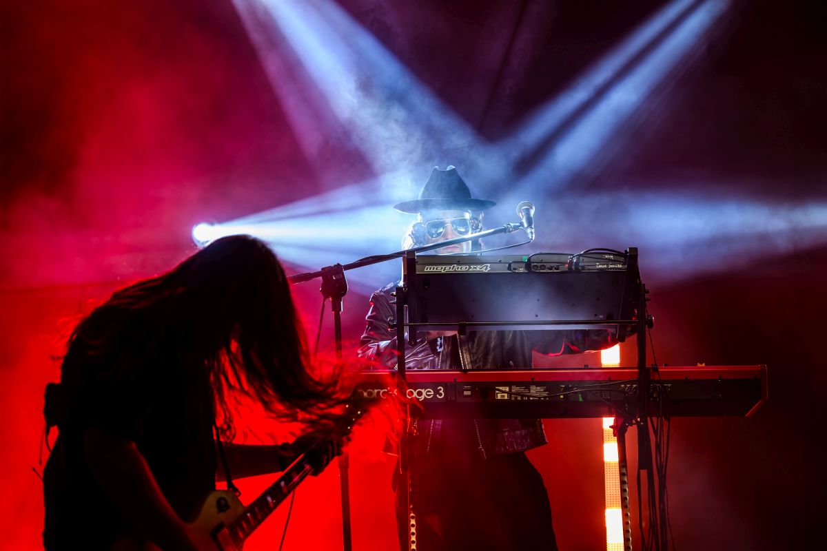 Muzyk gra na keyboardzie, w tle gitara. Dominują czerwone i niebieskie światła. Scena koncertowa pełna dymu i dramatyzmu.
