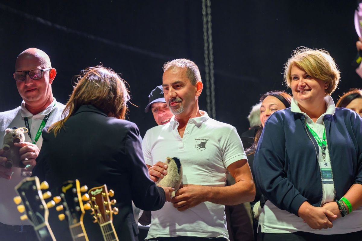 Na scenie stoi grupa osób w formalnych strojach, jedna wręcza pamiątkę. Tło czarne, z widocznymi gitarami. Kluczowe są wyrazy twarzy i interakcje.