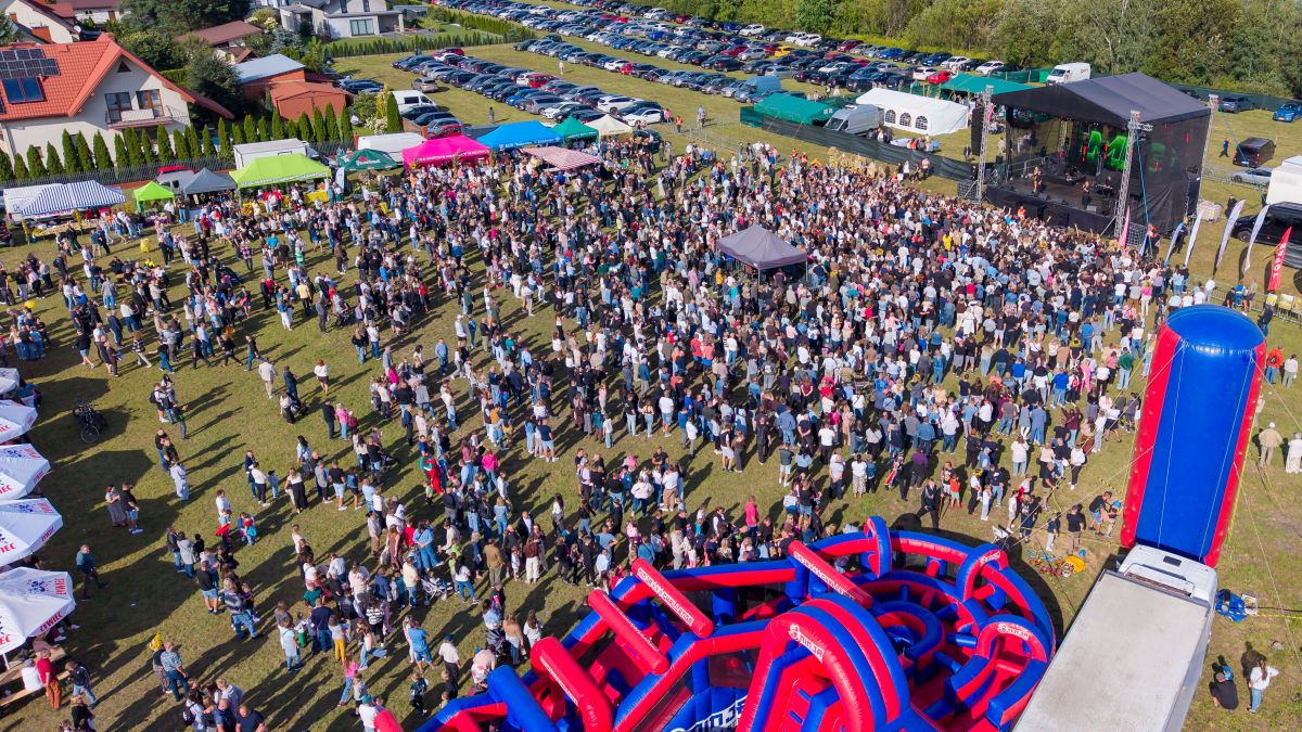 Zdjęcie przedstawia tłum ludzi na zewnątrz, uczestniczących w festiwalu. W tle scena, kolorowe namioty i zaparkowane samochody. Dominują zielone i niebieskie odcienie trawy i nieba.