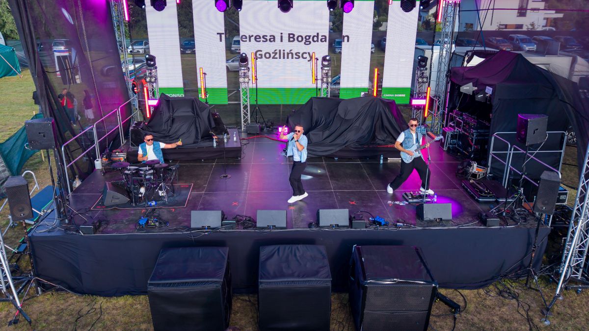 Trzech muzyków na scenie gra na instrumentach, ubrani w dżinsowe kamizelki. Scena jest oświetlona fioletowymi światłami. W tle widać duży ekran z napisami.