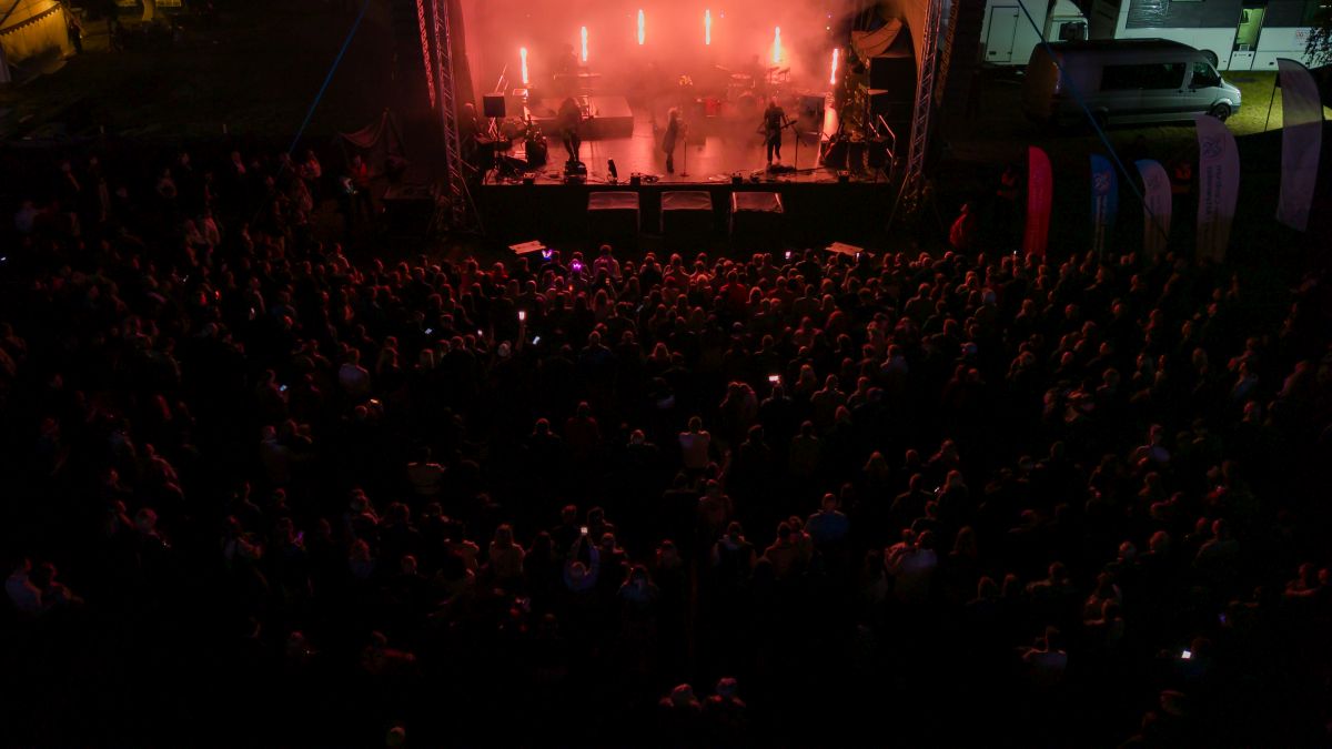 Koncert nocą: scena oświetlona czerwonym światłem, tłum ludzi na pierwszym planie. W tle ciemne niebo i namioty.