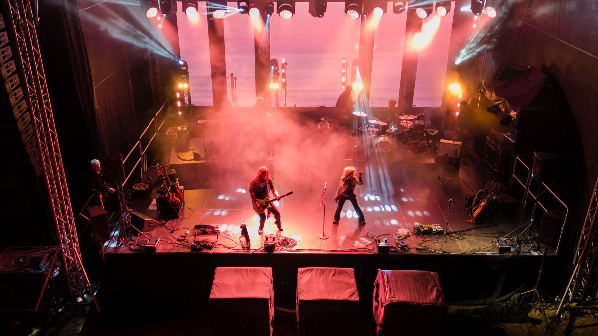 Scena koncertowa z muzykami w dymie i jasnych światłach. Przeważają kolory czerwony i pomarańczowy. Tło to ciemna przestrzeń z rampami oświetleniowymi.