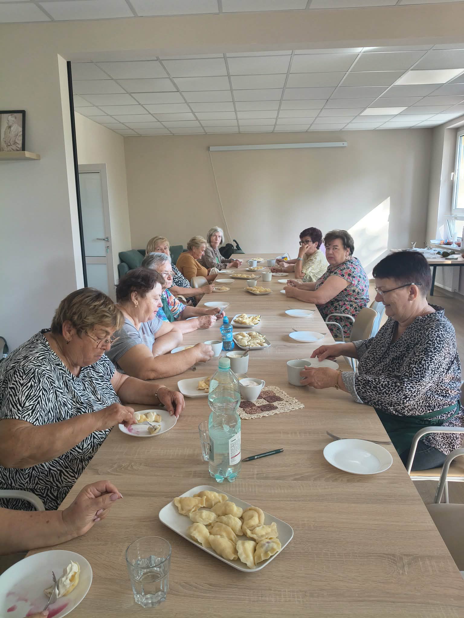 Na zdjęciu starsze kobiety siedzą przy długim drewnianym stole, jedząc pierogi. Stół jest nakryty białymi talerzami i szklanką z wodą. W tle jasne ściany i okno wpuszczające światło słoneczne.