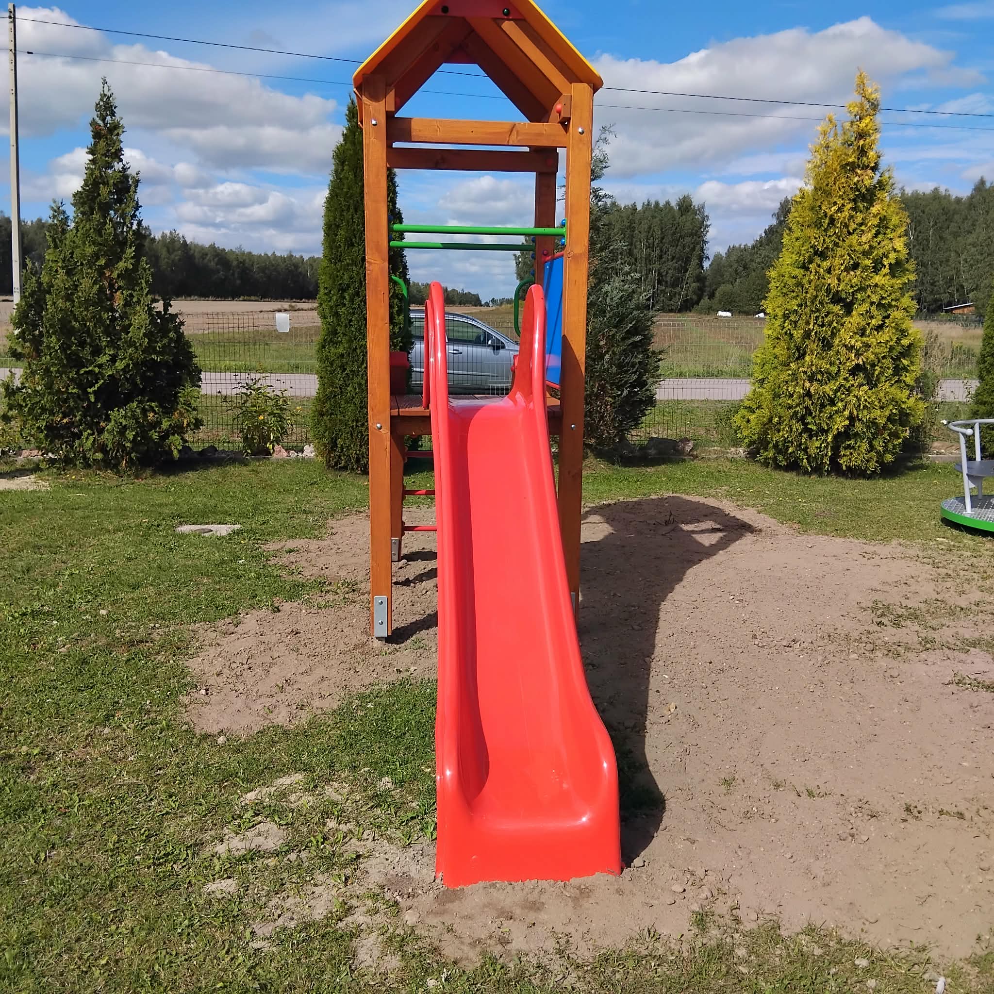 Czerwona zjeżdżalnia na placu zabaw z drewnianą konstrukcją, otoczona zielenią. W tle widać drzewa i błękitne niebo. Obok stoi zaparkowany samochód.