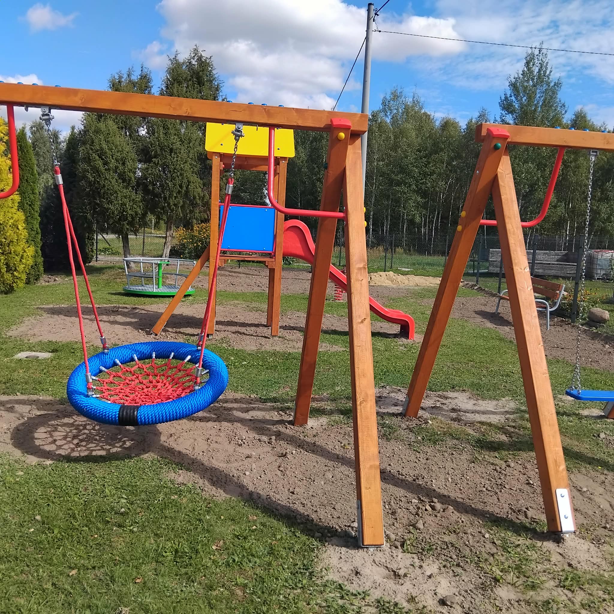 Plac zabaw z drewnianymi huśtawkami i kolorową zjeżdżalnią. Tło to trawnik, drzewa i błękitne niebo z białymi chmurami. Kolory są żywe: czerwony, niebieski i żółty.