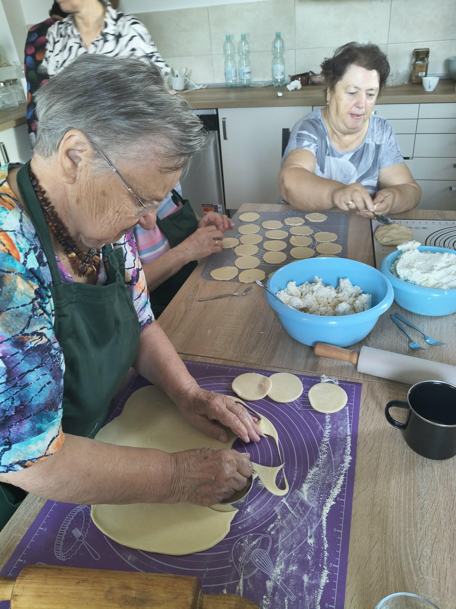 Kilka osób przygotowuje pierogi w kuchni, wykrawając ciasto. Stół jest drewniany, a miski niebieskie. W tle widać szafki i butelki.