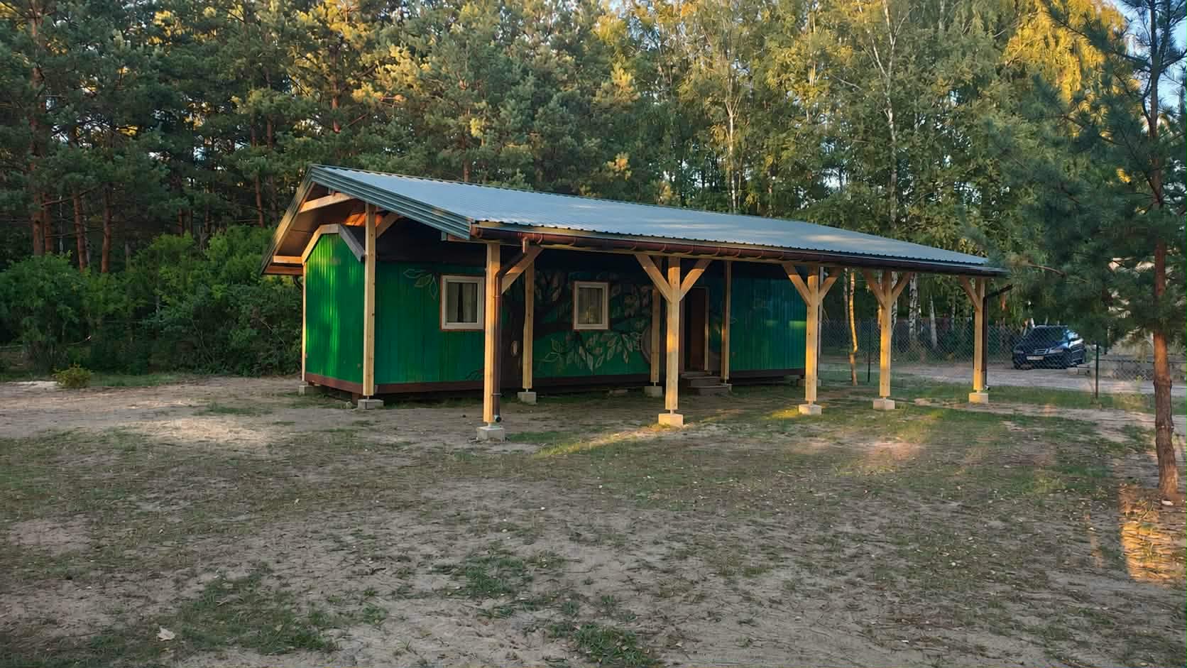 Zielona drewniana altana z dwuspadowym dachem stoi na tle leśnym. Teren wokół jest piaszczysty, a w tle widoczny jest zaparkowany samochód. Dookoła są drzewa i krzewy.