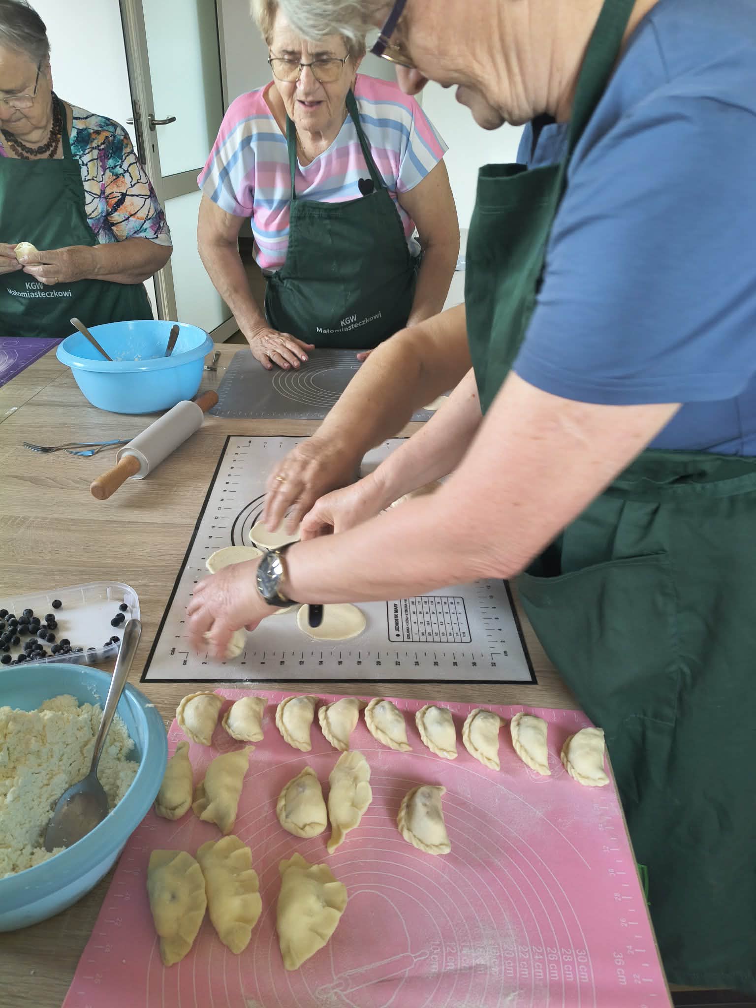 Trzy kobiety w zielonych fartuchach lepią pierogi przy drewnianym stole. Na stole są maty, wałek, miska z serem i jagody. Tło jest jasne, z białymi ścianami.