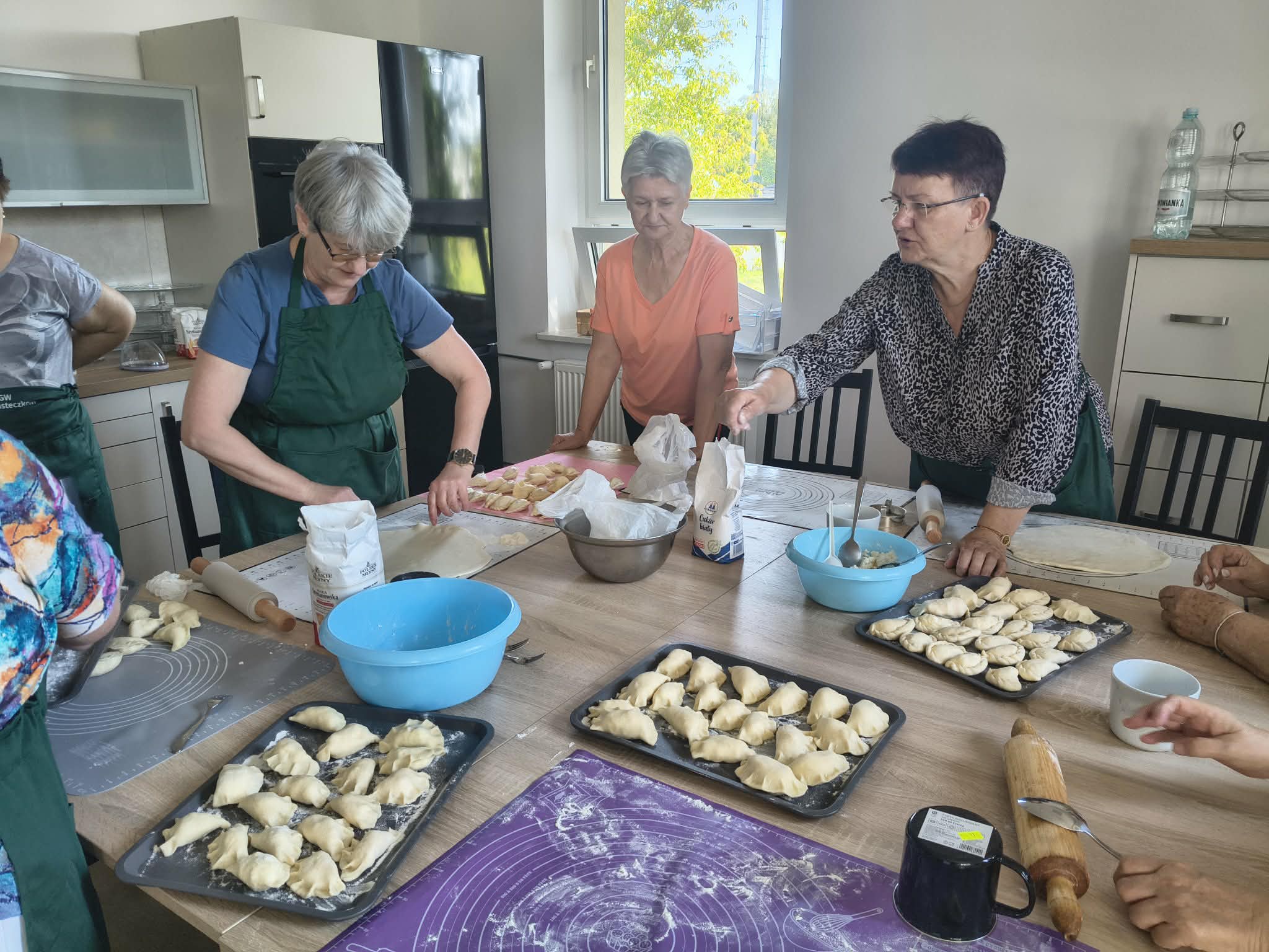 Grupa osób robi pierogi w kuchni, na drewnianym stole są patelnie z pierogami. Osoby mają na sobie zielone fartuchy, w tle jasne szafki i okno.
