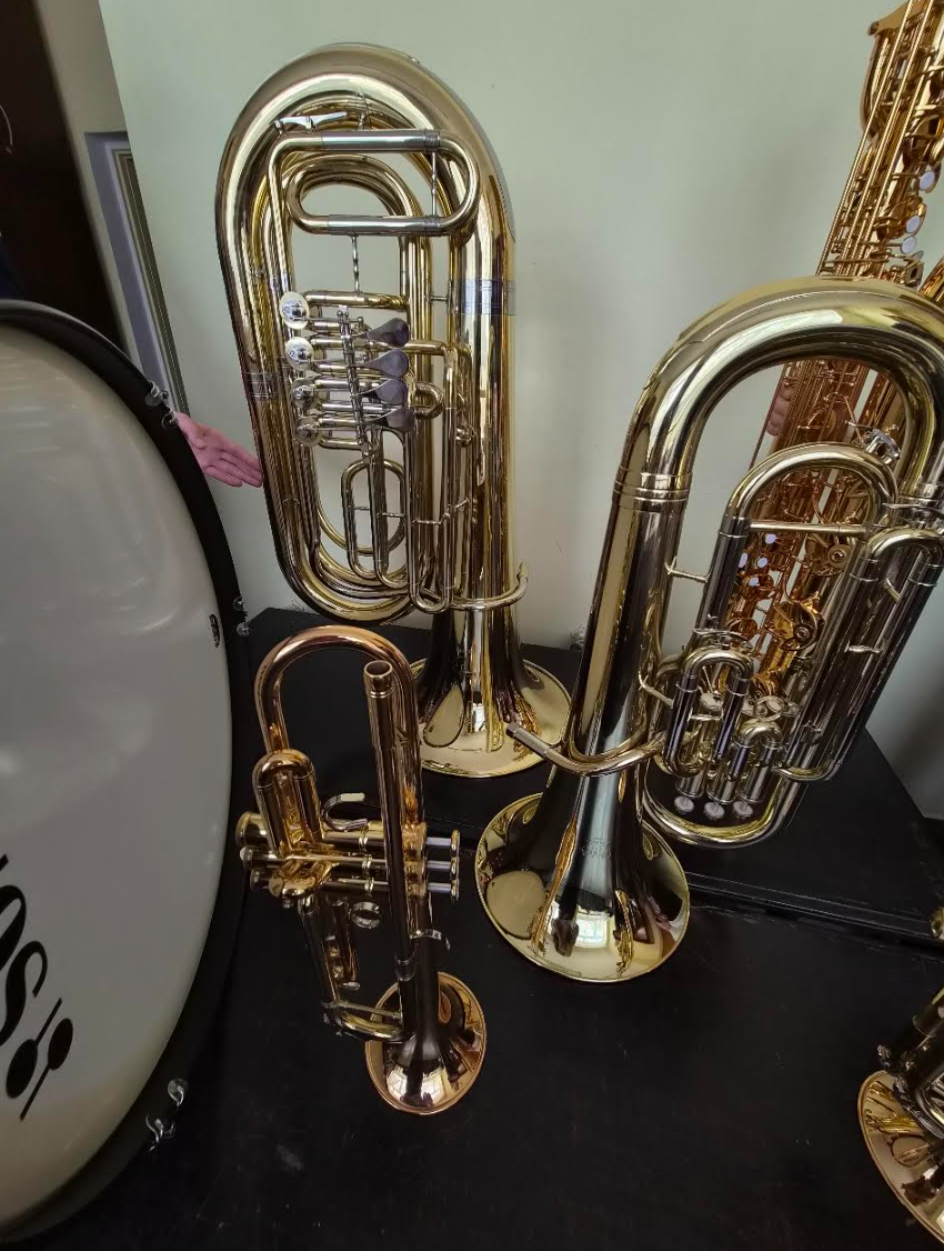 Złote instrumenty dęte stoją na ciemnym tle. Po lewej widoczny bęben. Jasna ściana tła kontrastuje z połyskiem metalu.