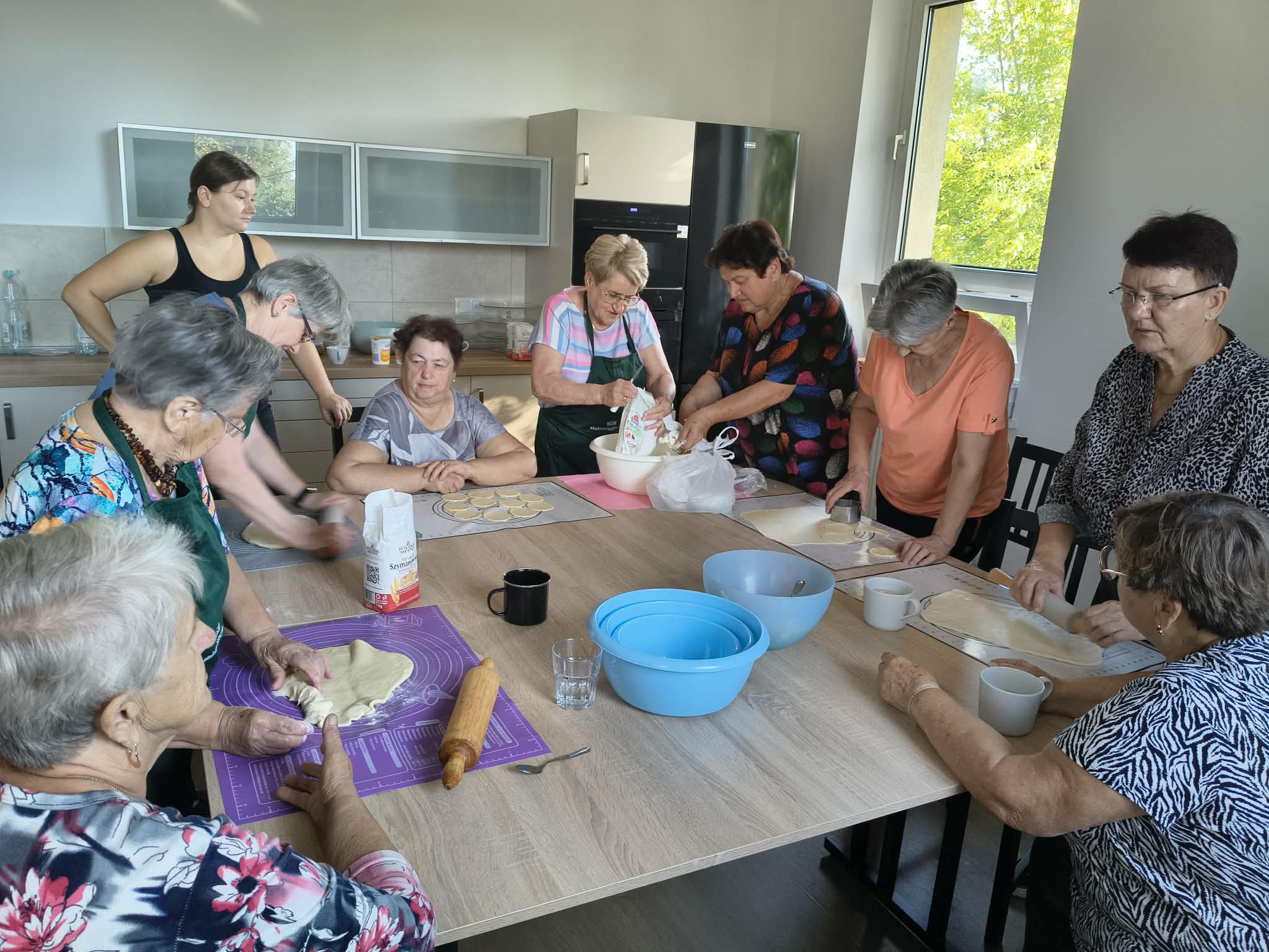 Grupa osób przy dużym stole pracuje nad przygotowywaniem ciasta. Wnętrze jasnej kuchni z drewnianymi szafkami. Na stole różne akcesoria kuchenne i miski.